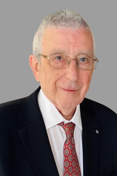 Portrait of a man with short grey hair who is wearing glasses, a red tie, white shirt and black suit. He is smiling.