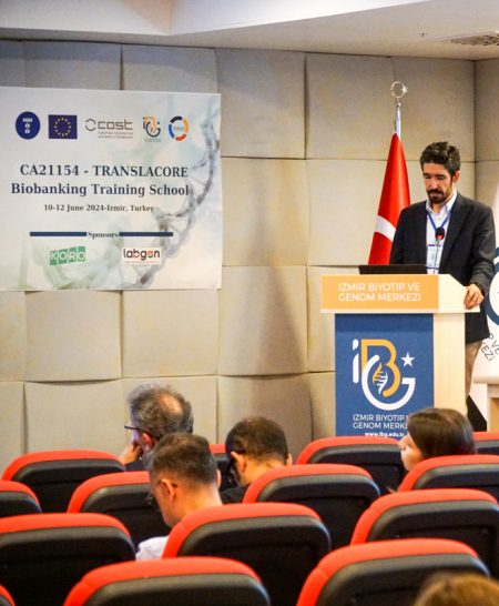 A man giving a presentation in a conference room in Izmir, Turkey, next to a poster promoting the current event, the Translacore training event.