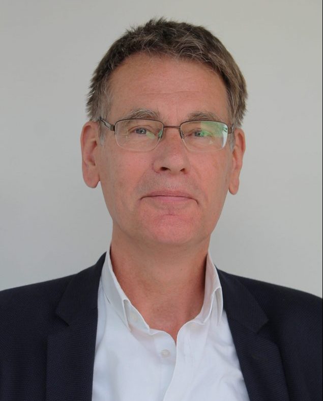 A white man with short fair hair and who wears glasses, a white shirt and black jacket, is smiling at the camera