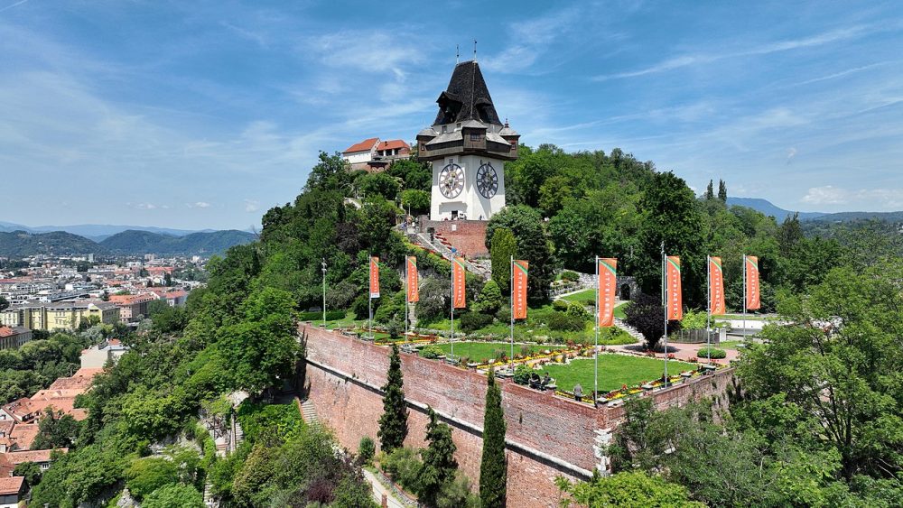 The Graz Clocktower is shown on top of the Graz Schlossberg, surrounded by trees. A small formal garden with red decorative flags is shown in front of the clock tower.