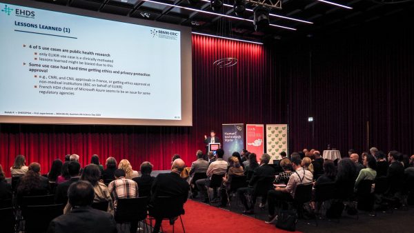 A white man wearing a grey suit and white shirt with short dark hair speaks in front of a projector screen about the European Health Data Space to an audience in a darkened auditorium with red curtains and carpet.