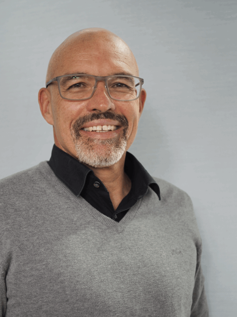 portrait of a white man with a shaved head and a beard. He is wearing glasses, a black shirt with a grey sweater over the top and he is smiling at the camera.