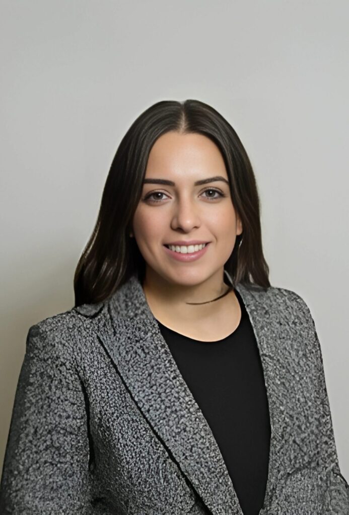 A woman with long dark hair in a professional, grey jacket, in front of a neutral grey background.